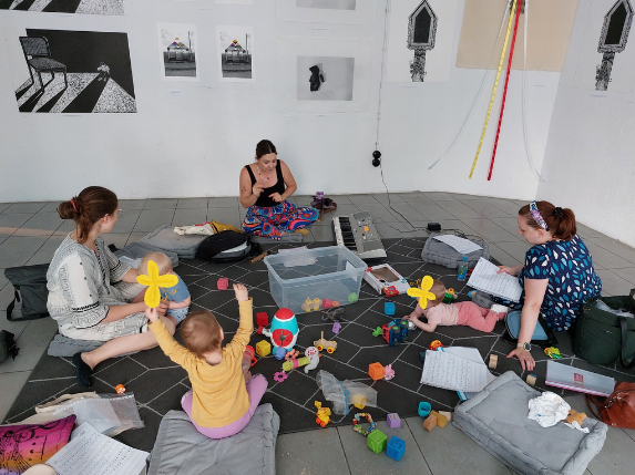 Mamy z dziećmi siedzące na podłodze. Wokół rozrzucone zabawki.