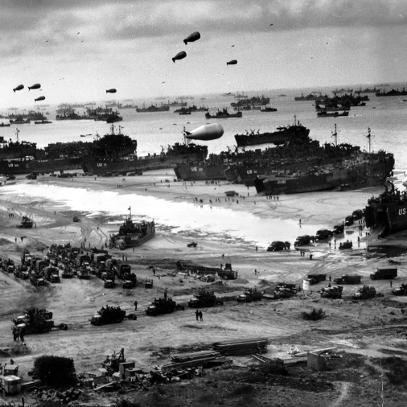 Biało-czarne zdjęcie plaży nad morzem wypełnionej samochodami, czołgami, wojskiem. Na wodzie liczne statki towarowe.
