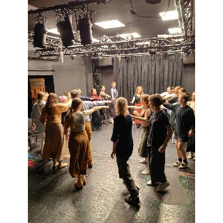 Grupa ludzi tańczy w kręgu na scenie. Prawa rękę mają wyciągnietą do środka.