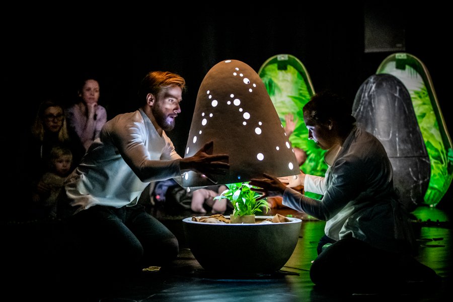 Dwoje ludzi otwiera świecącą kapsułę w której znajduje się roślinka. W tle można zaobserwować inne osoby które patrzą na całość z zaciekawieniem.