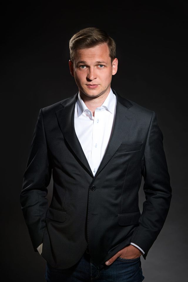 Na zdjęciu widoczny jest mężczyzna ubrany w elegancki czarny garnitur i białą koszulę. Stoi przed ciemnym tłem, które podkreśla jego sylwetkę. Mężczyzna ma krótkie jasne włosy i patrzy bezpośrednio w kamerę z lekkim uśmiechem. Ogólny ton fotografii tworzy wrażenie profesjonalizmu i pewności siebie.