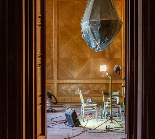 Przez otwarte drzwi widać fragment odnawianego pokoju. Na ścianach zabytkowa boazeria, żyrandol jest owinięty folią ochronną, a na podłodze stoją dwa krzesła, lampa i sporo kabli.