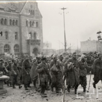 Czarno-biała fotografia idącej grupy mężczyzn. Za ich plecami budynek Zamku Cesarskiego.