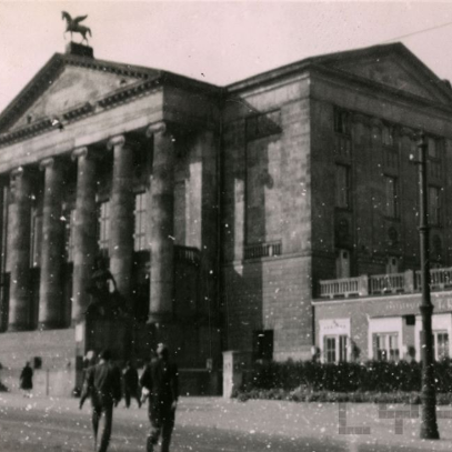 Czarno-białe zdjęcie Teatru Wielkiego w Poznaniu od frontu. Ulicą spacerują ludzie. Fotografia jest ziarnista, złej jakości.