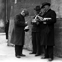 Czarno-białe zdjęcie trzech mężczyn w płaszczach i nakryciach głowy. Stoją na ulicy, w rękach trzymają książki.
