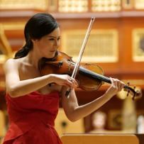 Czarnowłosa Azjatka gra na skrzypcach, na twarzy ma skupienie. Jest ubrana w czerwoną suknię z falbanami.
