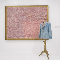 Duży obraz w ramie przypominający fakturą różowy materiał wisi na ścianie galerii. Przed obrazem stoi manekin krawiecki ubrany w dżinsową marynarkę.