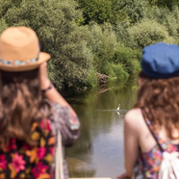 Dwie kobiety stojące tyłem patrzą na rzekę. Na wodzie widać czaplę, dookoła rzeki bujną roślinność.