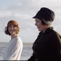 Dwie kobiety w kapeluszach, młodsza i starsza, spacerują brzegiem morza.