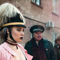 Dziewczyna w mocnym, mrocznym makijażu stoi na ulicy w górniczej czapce. Za nią dwaj mężczyźni, również w przebraniach.