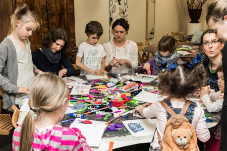 Opiekunowie z dziećmi na zajęciach przy stole pełnym kartek i kolorowych papierów.