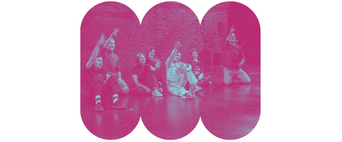 Grupa młodzieży siedzi na podłodze w sali gimnastycznej z prowadzącym zajęcia. Kadr na różowym tle.