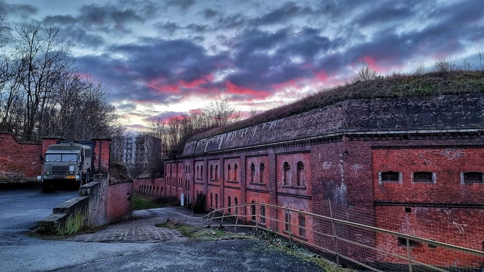 Budynki fortyfikacji z czerwonej cegły. Przy nich droga, a na niej samochód wojskowy.