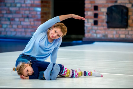 Kobieta siedzi z wyprostowanymi nogami na podłodze. Mała dziewczynka leży w poprzek na jej nogach.