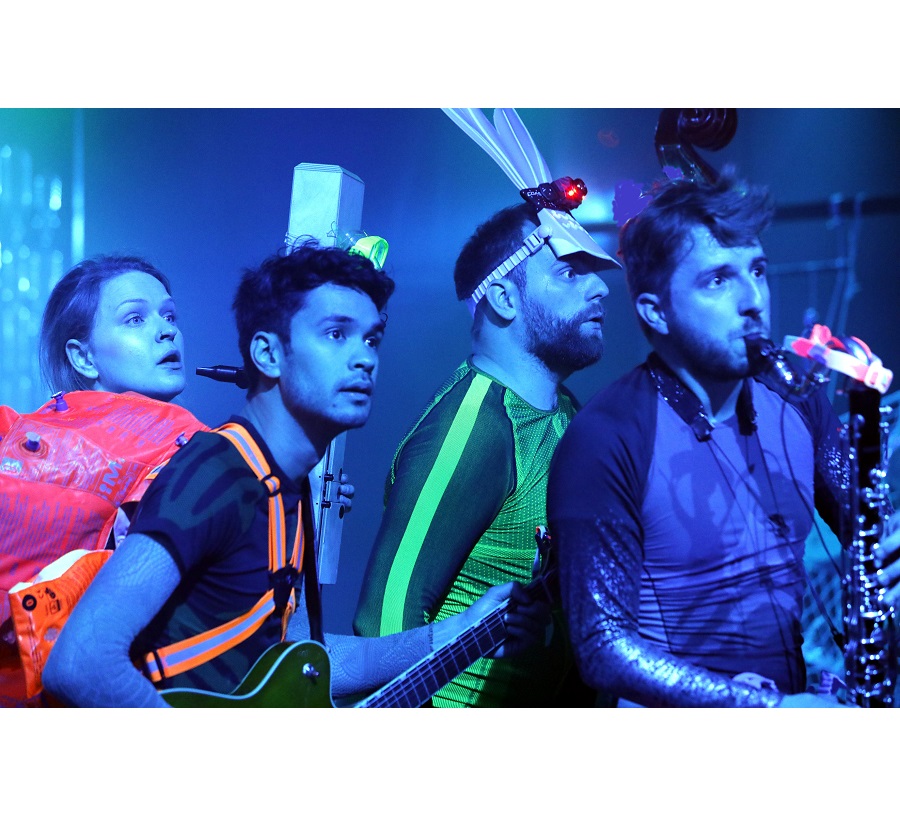 Na zdjęciu czworo młodych osób, stoją jeden za drugim, patrzą w tym samym kierunku, w rękach trzymają różne instrumenty muzyczne.