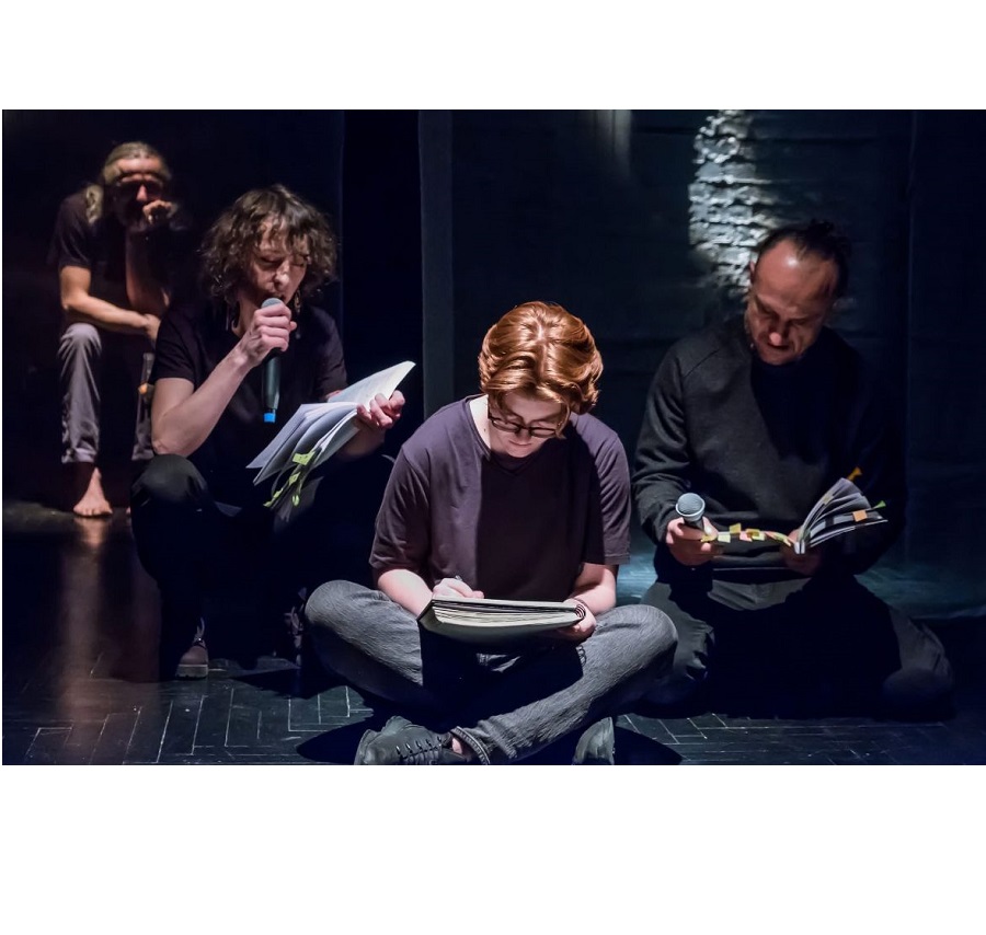 Kilku mężczyzn siedzi na scenie, każdy z nich trzyma przed sobą otwartą książkę. Jeden z mężczyzn patrząc na otwartą książkę czyta do mikrofonu.