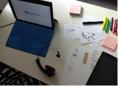 Laptop na stole, obok słuchawki, kartki, tablet, pisaki.