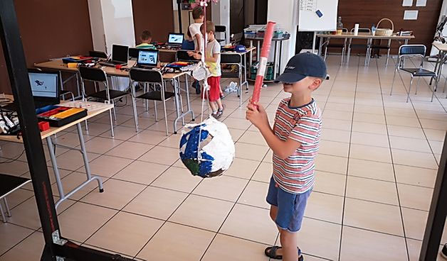 Sala, stoliki i krzesła. Na stołach laptopy. N apierwzym planie chłopiec z akrytymi czapka oczami z piankowa rurka próbuje trafić w zawieszona piłkę. Dalej 3 dzieci przy stolikach.