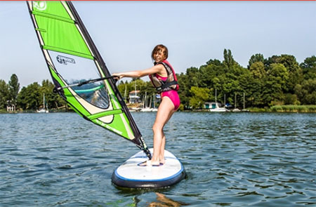 Dziewczyna w rózowym stroju kapielowym na desce windsurfingowej na jeziorze. Zielony żaiel.