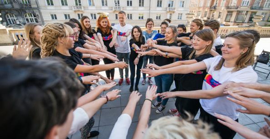 Młodzież stoi na placu w kręgu z wyciągniętymi rękoma do środka. W tle budynki.
