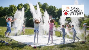 Fotografia przedstawia dzieci rzucające białym proszkiem ponad białym materiałem rozłożonym na trawie. Dodatkowo na fotografię nałożono logo 21. edycji Letniego Pogotowia Sztuki.