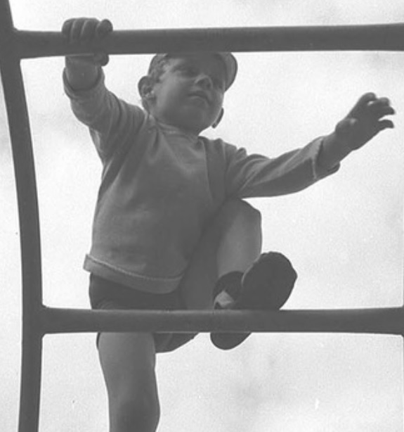 Czarno-biało fotografia przedstawiająca chłopca, który wspina się po drabinie. Na nogach ma wysokie podkolanówki. Nad nim widać zachmurzone niebo.