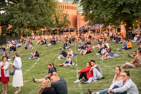 W parku ludzie siedzą na trawie w małych grupkach, najczęściej w parach i najprawdopodobniej słuchają koncertu.