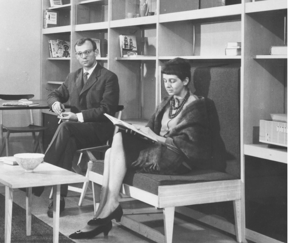 Na czarno-białej fotografii widzimy dwoje ludzi na tle dużego prostego regału. Kobieta znajdująca się bliżej siedzi na drewnianej tapicerowanej ławce będącej częścią regału. Przed nią stoi niewielki, prosty stolik kawowy, a za nią, na krześle, siedzi mężczyzna.