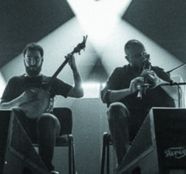 Czarno-białe zdjęcie wykonane zostało podczas koncertu, spod sceny. W centralnej jego części siedzi dwóch muzyków - jeden gra na banjo, drugi na dudach. Po prawej i po lewej ich stronie, nieco w głębi, znajdują się zastawione sprzętem elektronicznym stoły przy których stoi kolejnych dwóch muzyków. W głębi sceny tańczą snopy scenicznych reflektorów.