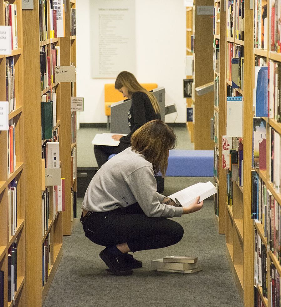 Fotografia dwóch kobiet kucających między regałami z książkami i czytającymi książki.