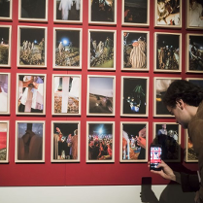 Fotografie z wydarzenia są oprawione w białe ramki i wiszą wszystkie (jest ich barszo dużo) na czerwonej ścianie. Stoi pod nimi zwiedzający mężczyzna, który jednej z fotografii robi zdjęcie telefonem.