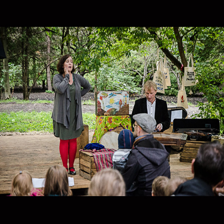 Dwoje aktorów, kobieta i mężczyzna, w trakcie spektaklu. Przed nimi grupa osób oglądających spektakl, w tle park i drzewa.