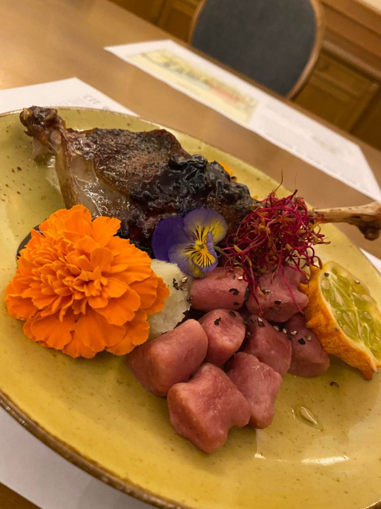 Zdjęcie, na talerzu noga gęsia, obok różowe kluseczki, kwiaty jadalne