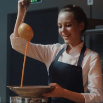 Główna bohaterka w uniformie i fartuchu przyrządza deser, patrzy z radością i apetytem na łyżkę, z której przelewa się do talerza pomarańczowy krem.