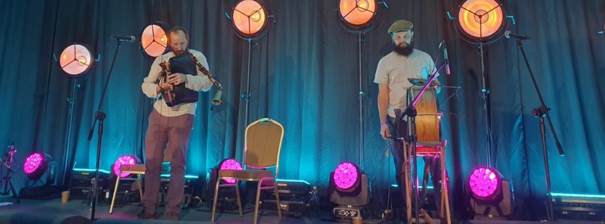 Dwóch mężczyzn na scenie. Jeden gra na dudach a drugi na bębnie. Na scenie stoją też lampy z pomarańczowym i różowym światłem. Z tyłu turkusowa kotara.