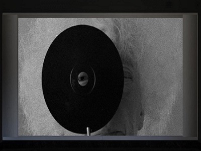 Czarno-biała grafika. Na pierwszym planie widać czarną okrągłą płytę z otworem w samym jej środku. Przez otwór spogląda ludzkie oko. W dalszej perspektywie widać, że za płytą stoi rezydentka.