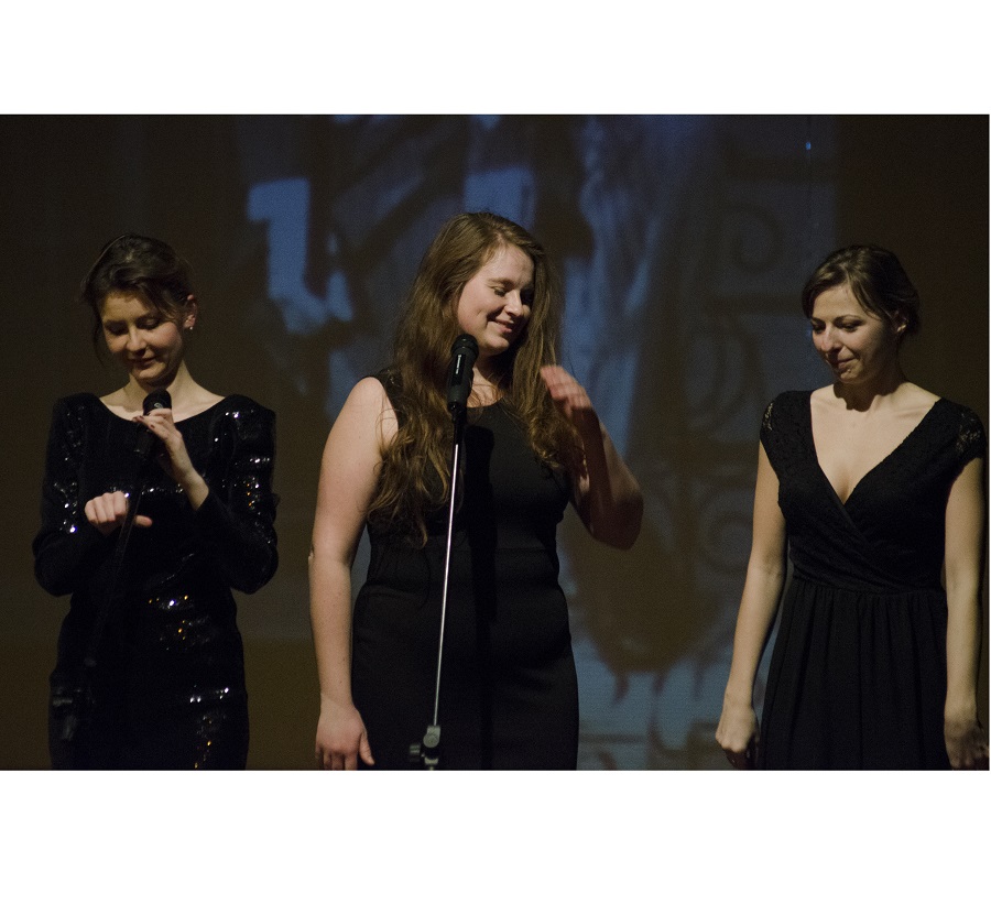 Zdjęcie przedstawia trzy kobiety, w czarnych sukienkach, stojące na scenie podczas koncertu. Przed nimi stoją mikrofony.