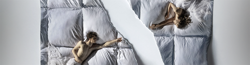 Kobieta i mężczyzna leżą na jasnej pościeli naprzeciwko siebie po skosie, wyciągając do siebie ręce.