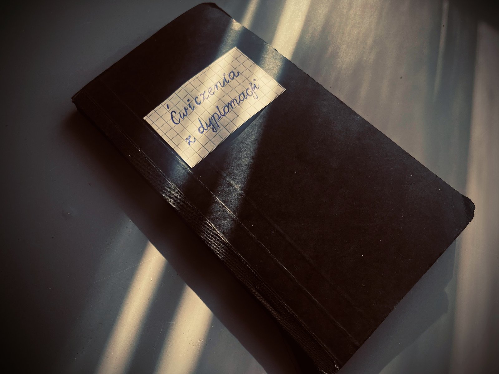 na zdjeciu widoczny czarny zeszyt, na okładce w gornej części naklejony prostokąt z kartki zeszytowej w kratkę, z napisem "Ćwiczenia z dyplomacji"