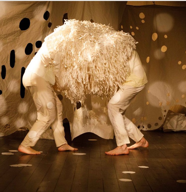 grafika przedstawia dwie bose, pochylone postaci stojące przodem do siebie, na głowach i barkach mają narzuconą narzutę z długimi frędzlami