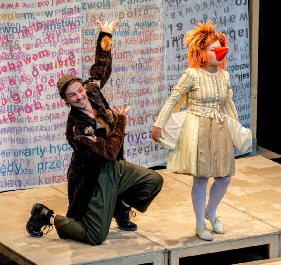 po lewej stronie zdjęcia klęczący na jedno kolano mężczyzna w czapce, ubrany w długi futrzany brązowy płaszcz, po prawej stojąca kobieta w sukience, rudej peruce i pomarańczowym doklejonym dziobie