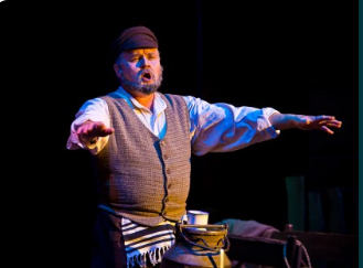 pośrodku zdjęcia mężczyzna siedzący z wyciągniętymi w bok obiema rękami, przed nim widoczna górna część dużej kanki na mleko, na niej kubek; mężczyzna śpiewa.