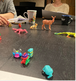 na zdjęciu widoczny stół z leżącymi kolorowymi figurkami zwierząt, za stołem widoczne fragmenty dwóch siedzących postaci