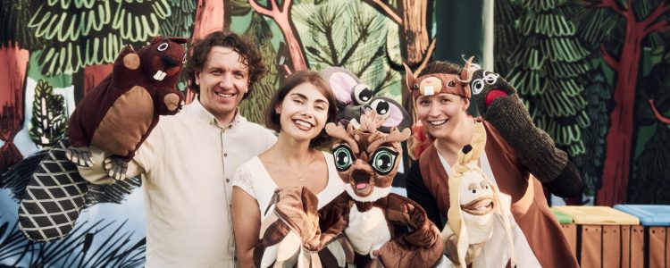 Zdjęcie trójki aktorów, którzy trzymają lalki - bohaterów spektaklu Grajmy w zielone. Aktorzy stoją na tle rysunkowego lasu.