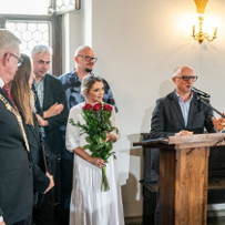 Grupa ludzi odbiera nagrodę w zabytkowym ratuszu. Z mównicy przemawia mężczyzna, kobieta obok niego trzyma bukiet róż. Po lewej stoi prezydent miasta w złotym łańcuchu na szyi.