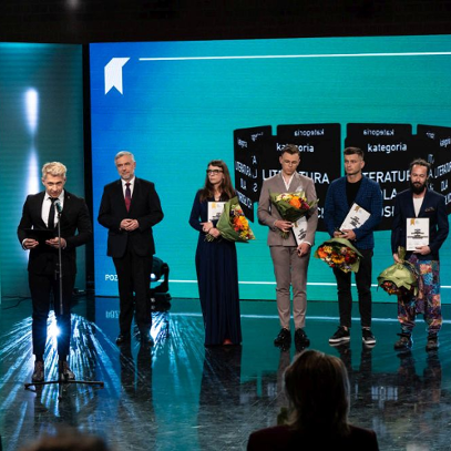 Grupa ludzi stoi na scenie trzymając dyplomy i bukiety kwiatów. Dwoje prowadzących galę wręczenia nagród przemawia do mikrofonów na statywach.