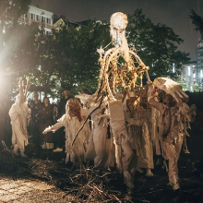 Grupa postaci zwierząt niesie na ramionach coś, co przypomina statuę uformowaną z ważnych dla nich przedmiotów.