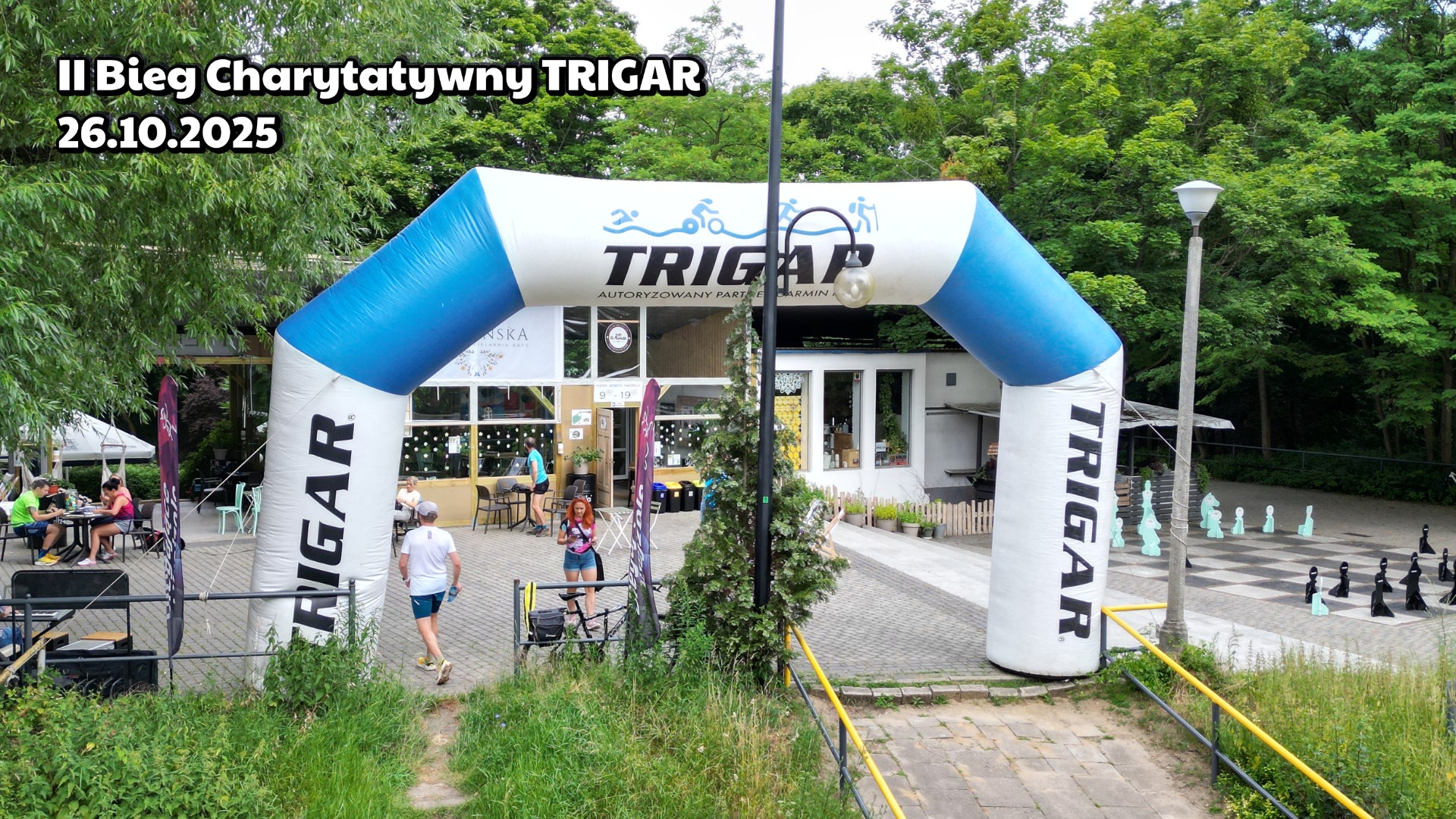 Meta biegu charytatywnego. W tle budynek kawiarni w środku parku. Po prawej duża szachownica i dużymi pionkami.