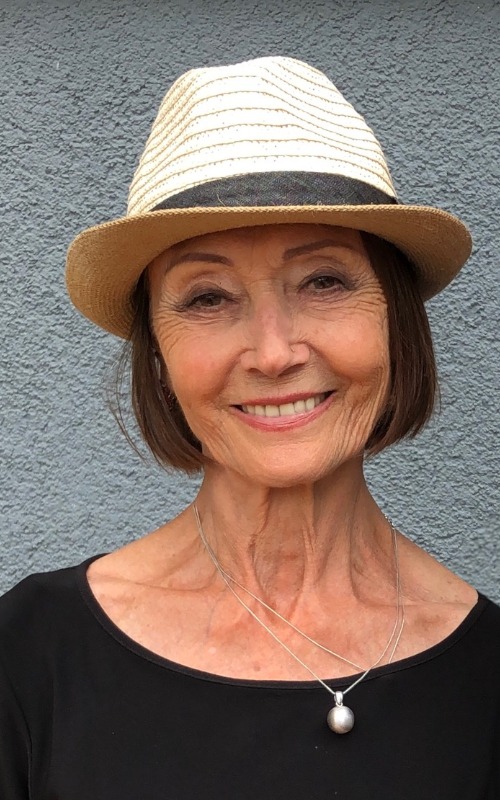 Ewa Wydyłło. Porter kobiety w średnim wieku. Kobieta ma krótkie, brązowe włosy, na głowie kapelusz. Patrzy przed siebie i szeroko się uśmiecha.