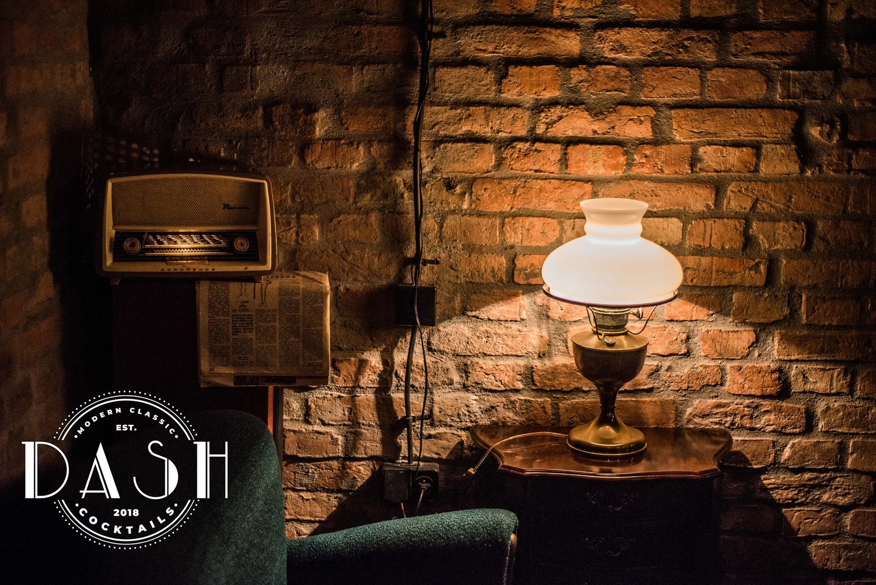 Stara miedziana lampa z ozdobnym kloszem stoi na stoliku. Oświetla zielony fotel, rozwieszoną gazetę i stare radio. W tle ceglana ściana.